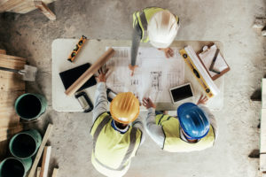 AERIAL VIEW OF THREE PEOPLE IN HARDHATS STANDING AROUND A TABLE THAT HAS CONSTRUCTION BLUEPRINTS LAID OUT ON IT.