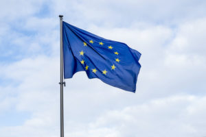 A EUROPEAN UNION FLAG WAVING IN THE WIND ATOP A FLAG POLE IN FRONT OF A CLOUDY, BLUE SKY.