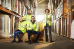 A GROUP OF YOUNG PEOPLE IN HIGH VIS VESTS STAND IN A WAREHOUSE AISLE, SMILING