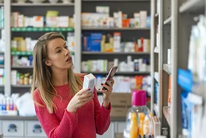 A WOMAN IN A SMALL PHARMACY HOLDS A MEDICATION IN ONE HAND AND HER CELL PHONE IN THE OTHER.