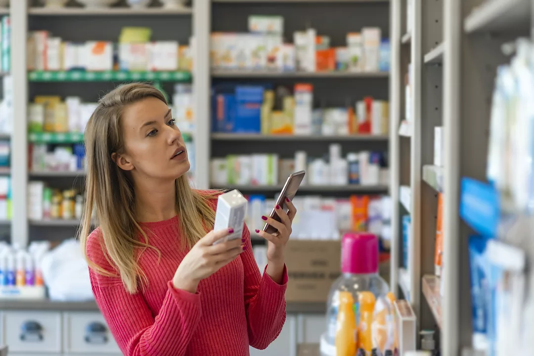 A WOMAN IN A SMALL PHARMACY HOLDS A MEDICATION IN ONE HAND AND HER CELL PHONE IN THE OTHER.