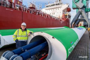 A MAN IN A HI-VIS JACKET AND HARDHAT LEANS ON TWO LARGE ECONOWIND SAILS NEXT TO A CHEMICAL TANKER