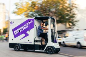 A WHITE AND PURPLE COVERED CARGO BIKE HAS THE FIN LOGO AND THE WORDS DELIVERING PARCELS NOT POLLUTION ON ITS SIDE