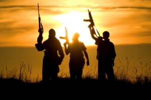 THREE FIGURES HOLDING ALOFT MACHINE GUNS ARE SILHOUETTED BY A SETTING SUN