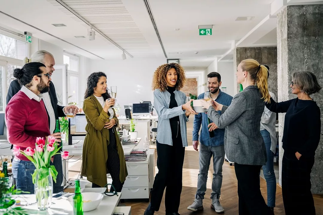 IN A GROUP OF PEOPLE STANDING IN AN OFFICE, ONE IS HANDING A SMALL BOX TO ANOTHER WHILE A THIRD DRINKS FROM A CHAMPAGNE GLASS