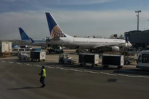 A WORKER IN HI-VIS VEST CROSSES THE TARMAC OF AN AIRPORT WITH TWO UNITED PLANES PARKED IN THE BACKGROUND