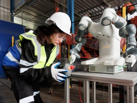 A WORKER IN A HARDHAT AND HI-VIS VEST HOLDS UP A MANUFACTURING COMPONENT TO A ROBOTIC ARM