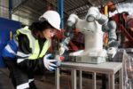 A WORKER IN A HARDHAT AND HI-VIS VEST HOLDS UP A MANUFACTURING COMPONENT TO A ROBOTIC ARM