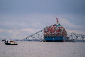 A HUGE CONTAINER SHIP SITS UNDER A COLLAPSED BRIDGE STRUCTURE
