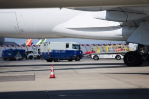 A TRUCK LABELED WITH THE BRINKS WORDING IS SEEN IN THE DISTANCE, UNDER THE BELLY OF A STATIONARY AIRCRAFT ON THE TARMAC OF AN AIRPORT