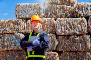 A WORKER IN A HARD HAT AND HI-VIS ACCENTED JACKET STANDS IN FRONT OF TOWERING STACKS OF BALES OF RECYCLING WASTE.