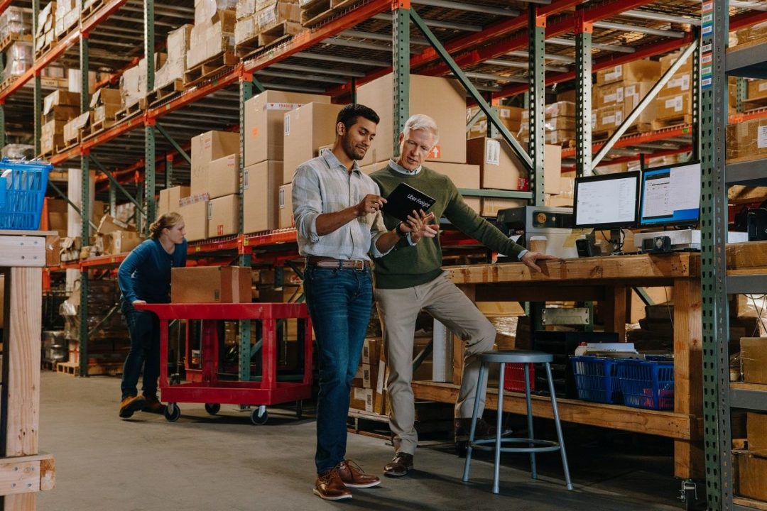 TWO WORKERS DISCUSS WHAT'S ON A TABLET COMPUTER IN A WAREHOUSE