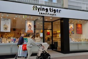 TWO PEDESTRIANS PUSH A SUITCASE AND STROLLER PAST A BRIGHT SHOP FRONT LABELED FLYING TIGER.
