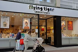 TWO PEDESTRIANS PUSH A SUITCASE AND STROLLER PAST A BRIGHT SHOP FRONT LABELED FLYING TIGER.