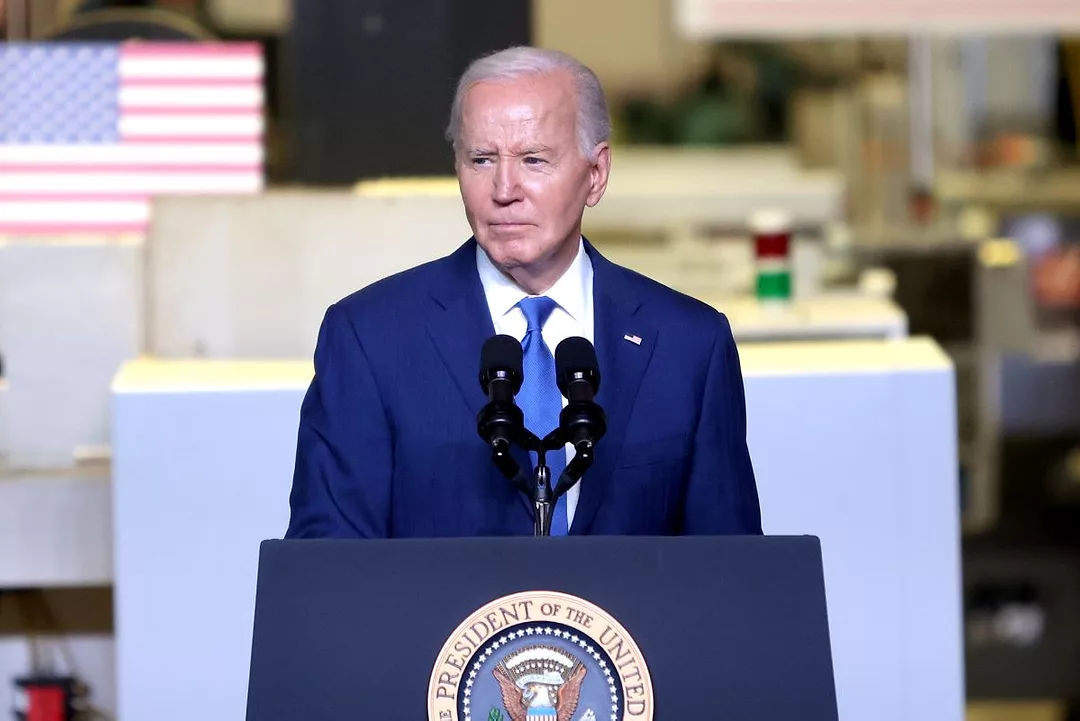 AN ELDERLY MAN IN A BLUE SUIT STANDS BEHIND A PODIUM LABELED PRESIDENT OF THE UNITED STATES