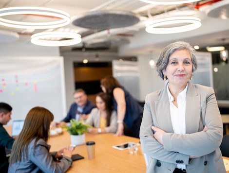 A GREY-HAIRED WOMAN IN A SHARP SUIT STANDS SMILING AT THE END OF A CONFERENCE TABLE WHERE OTHERS ARE GATHERED