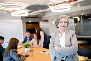 A GREY-HAIRED WOMAN IN A SHARP SUIT STANDS SMILING AT THE END OF A CONFERENCE TABLE WHERE OTHERS ARE GATHERED