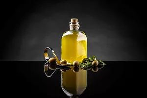 A BOTTLE OF OLIVE OIL STANDS SURROUNDED BY OLIVES ON A BEAUTIFUL DARK REFLECTIVE SURFACE