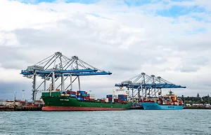 A green container ship unloading cargo at a port, with a pair of blue cranes above it