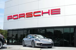 A grey sports car parked outside of a Porsche dealership, with the carmaker's brand written in red on the outside of the building