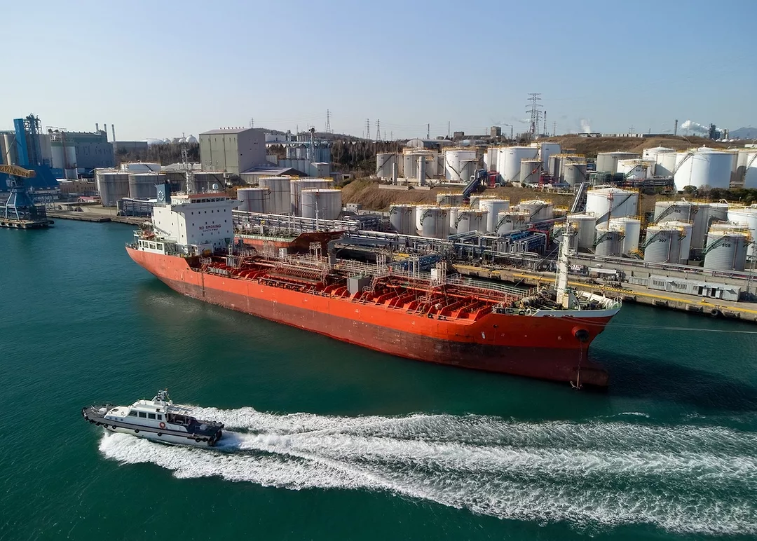 A red oil tanker along a shoreline full of white cylindrical silos
