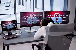 AN OFFICE WORKER HOLDS HER HEAD IN HER HANDS IN FRONT OF 3 SCREENS THAT ALL BEAR THE MESSAGE: YOUR PERSONA FILES ARE ENCRYPTED