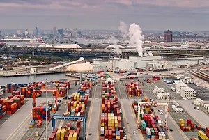 Rows of stacked, multi-colored shipping containers in a shipyard, with a collection of white industrial buildings in the background