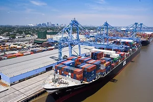 A black container ship docked at a port, under two large blue shipping cranes.