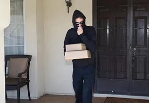 A man in a black hoodie pulled over his face, holding two packages on a front porch