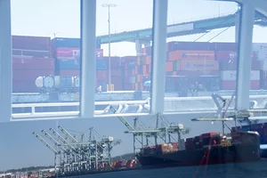 A window looking out at a container ship docked at a shoreline next to blue shipping cranes and stacks of containers