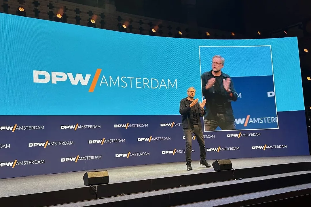 A MAN IN CASUAL BLACK CLOTHES ADDRESSES AN AUDIENCE FROM A STAGE THAT BEARS A DPW AMSTERDAM BACKDROP