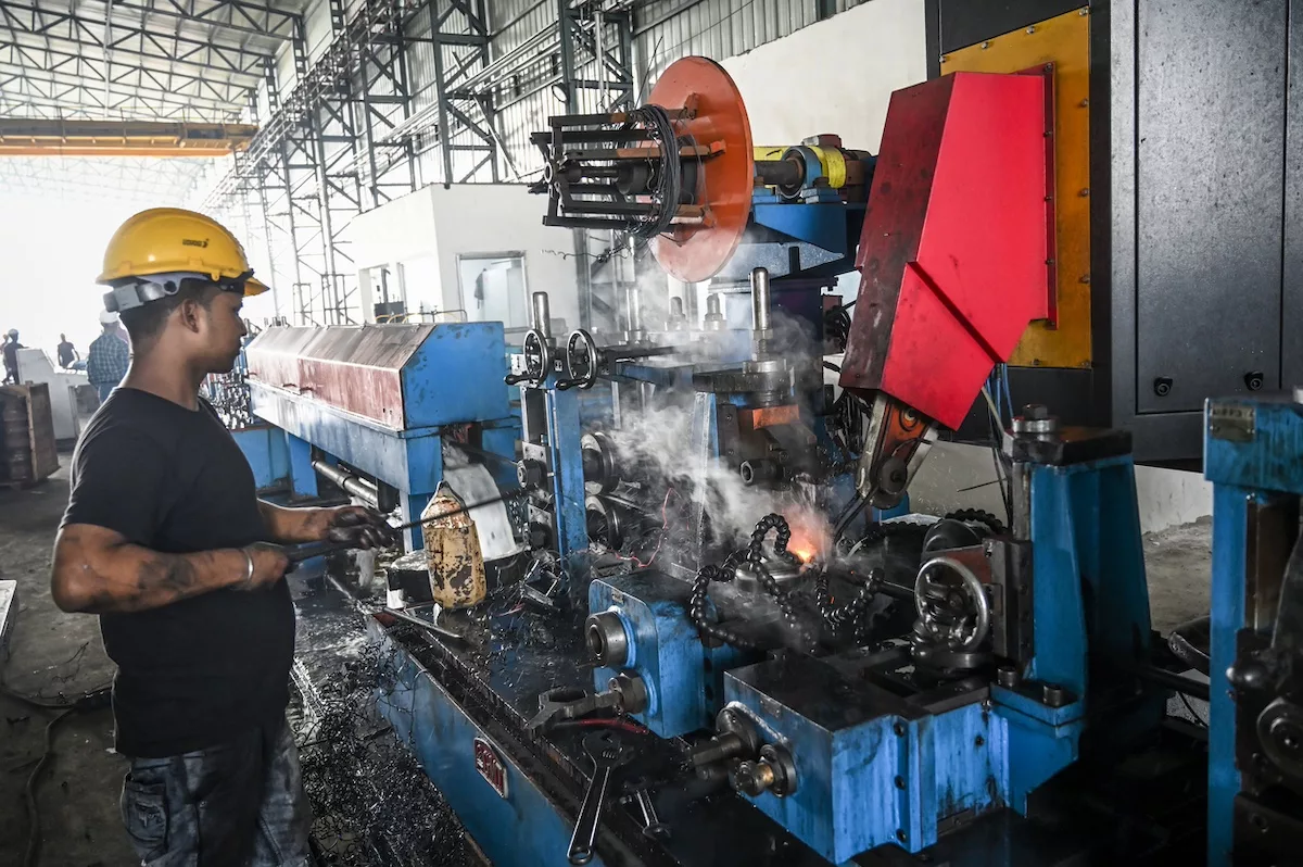 Siyah tişört ve sarı baret giyen bir adam, fabrika montaj hattındaki büyük mavi endüstriyel makinenin önünde duruyor