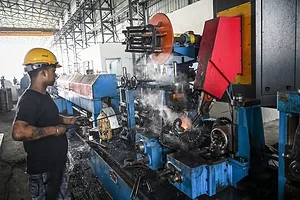 A man in a black t-shirt and a yellow hard hat standing in front of a large blue industrial machine on a factory assembly line.
