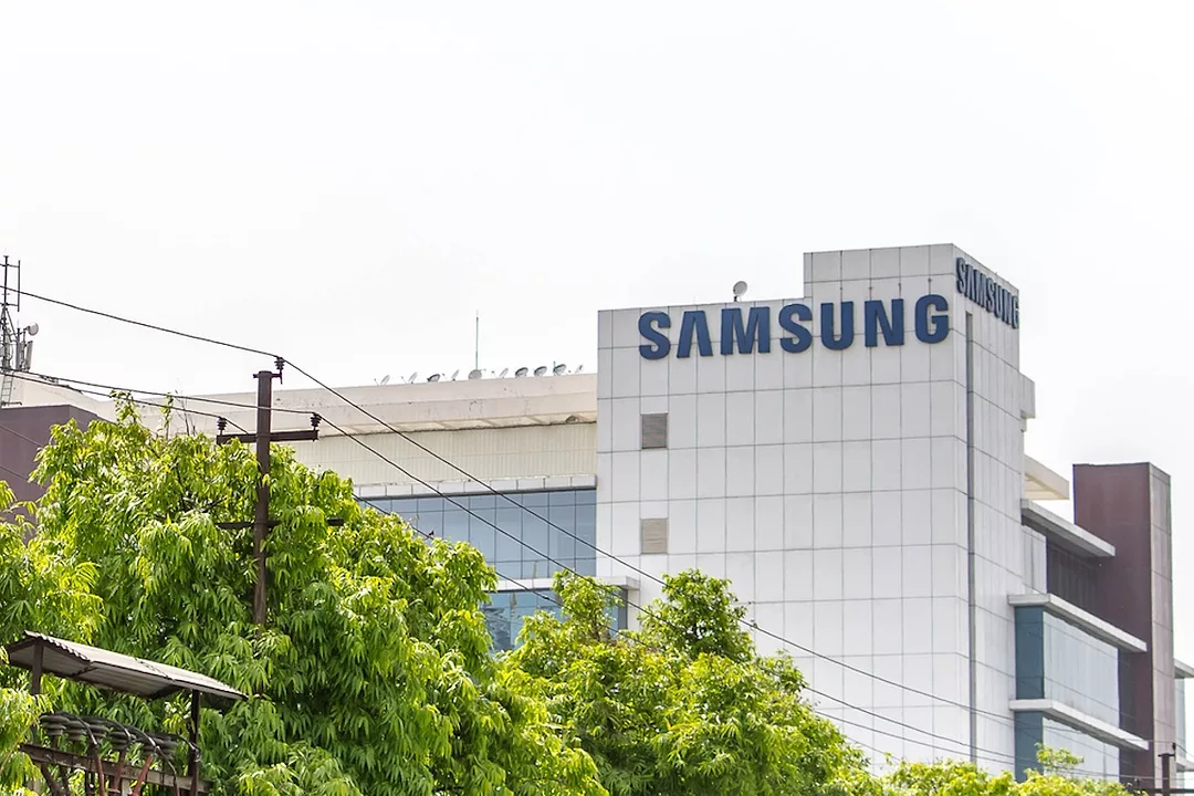 A white office building with "SAMSUNG" written in large blue letters at the top right, with a row of trees in the foreground