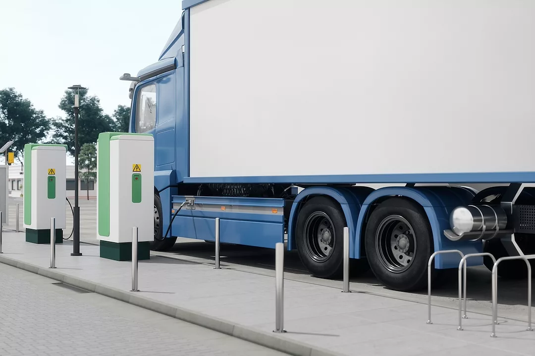 A blue semi-truck with a white cab, parked along a curb next to two white and green charging boxes.
