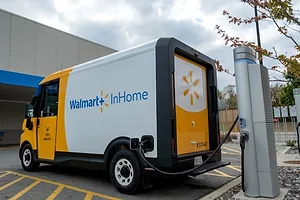 A yellow and white delivery van plugged into an electric charger, with "Walmart InHome" written along the side in blue letters. 