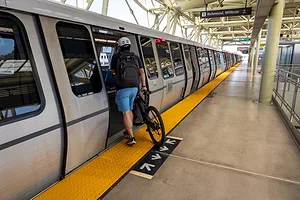 A PERSON IN BLUE SHORTS PUSHES A BICYCLE ON BOARD A LIGHT RAIL CAR