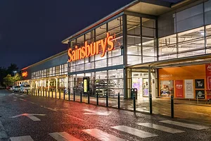 The front of a grocery store with large glass windows, orange walls, and "Sainsbury's" written in large orange neon letters across the top above the entrance.