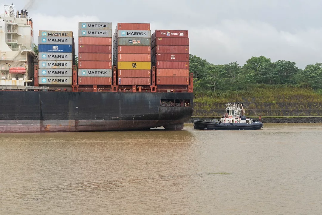 A black container ship seen from the side, with four large stacks of red and grey shipping containers on the front end.