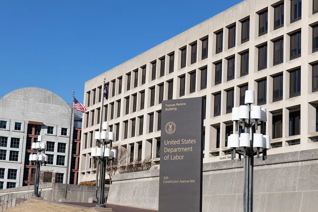 A large white concrete building with rows of black windows, with a black plinth out front that reads "United States Department of Labor" in gold lettering