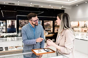 A MAN AND A WOMAN SMILE AND TALK OVER ITEMS OF JEWELRY ON TOP OF A DISPLAY CASE IN A STORE
