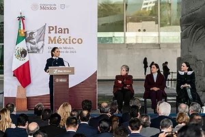 A WOMAN IN A SUIT, HER HAIR TIED BACK, MAKES AN ADDRESS TO AN OUTDOOR AUDIENCE FROM A PODIUM IN FRONT OF THE WORDS PLAN MEXICO AND A MEXICAN FLAG