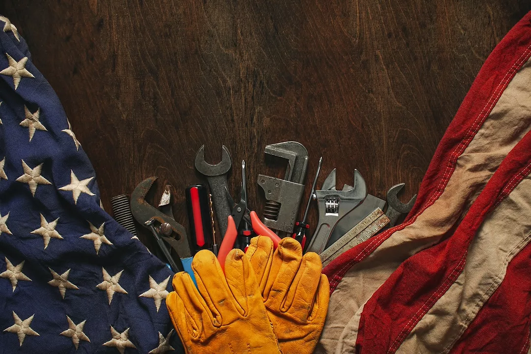 Yellow work gloves set on a brown wood table on top of an American flag, set beneath a row of wrenches and other tools.