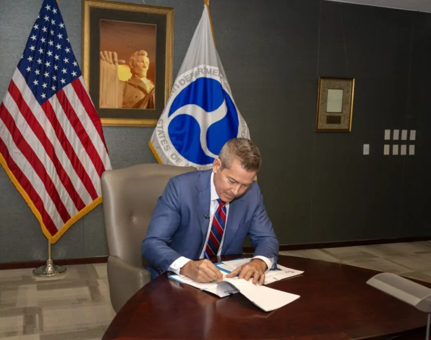 A MAN IN A BLUE SUIT AND RED TIE SITS AT A WOODEN TABLE, SIGNING PAPERS, IN FRONT OF A US FLAG AND A PORTRAIT OF PRESIDENT LINCOLN