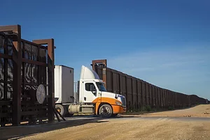 An orange and white semi-truck parked at the open gate of a long brown grated wall spanning the U.S.-Mexico border