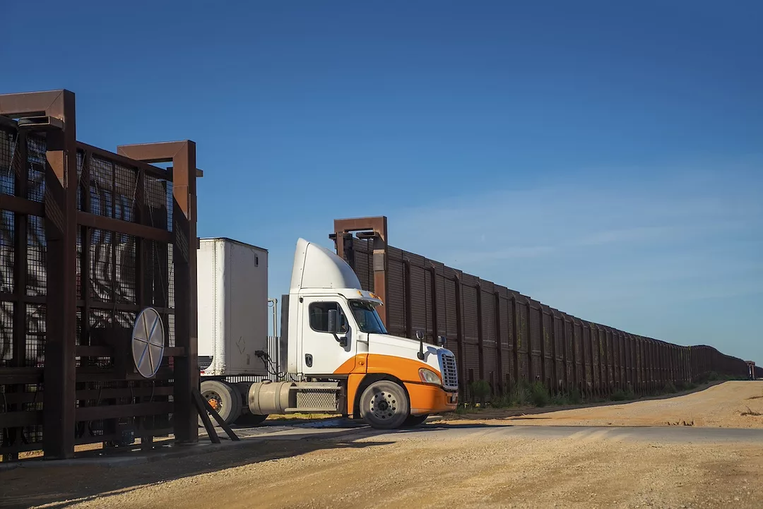 An orange and white semi-truck parked at the open gate of a long brown grated wall spanning the U.S.-Mexico border
