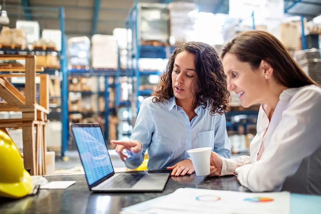 Two women sitting in front of a laptop in a warehouse, inside of a warehouse with rows of metal blue shelving stacked with boxes in the background