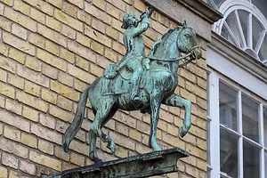 An oxidised bronze sign of a post boy or man on horseback attached to the wall of a building.