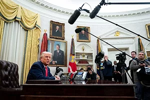 A MAN IN A BLUE SUIT AND RED TIE SITS AT A DESK IN FRONT OF A CURVED WALL, SURROUDED BY PEOPLE WITH LONG MICROPHONES