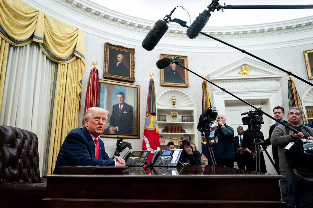 A MAN IN A BLUE SUIT AND RED TIE SITS AT A DESK IN FRONT OF A CURVED WALL, SURROUDED BY PEOPLE WITH LONG MICROPHONES